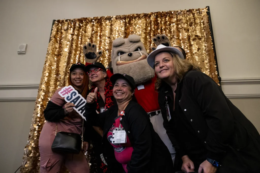 Hairy Dawg poses in front of a gold photo backdrop with smiling UGA fans.