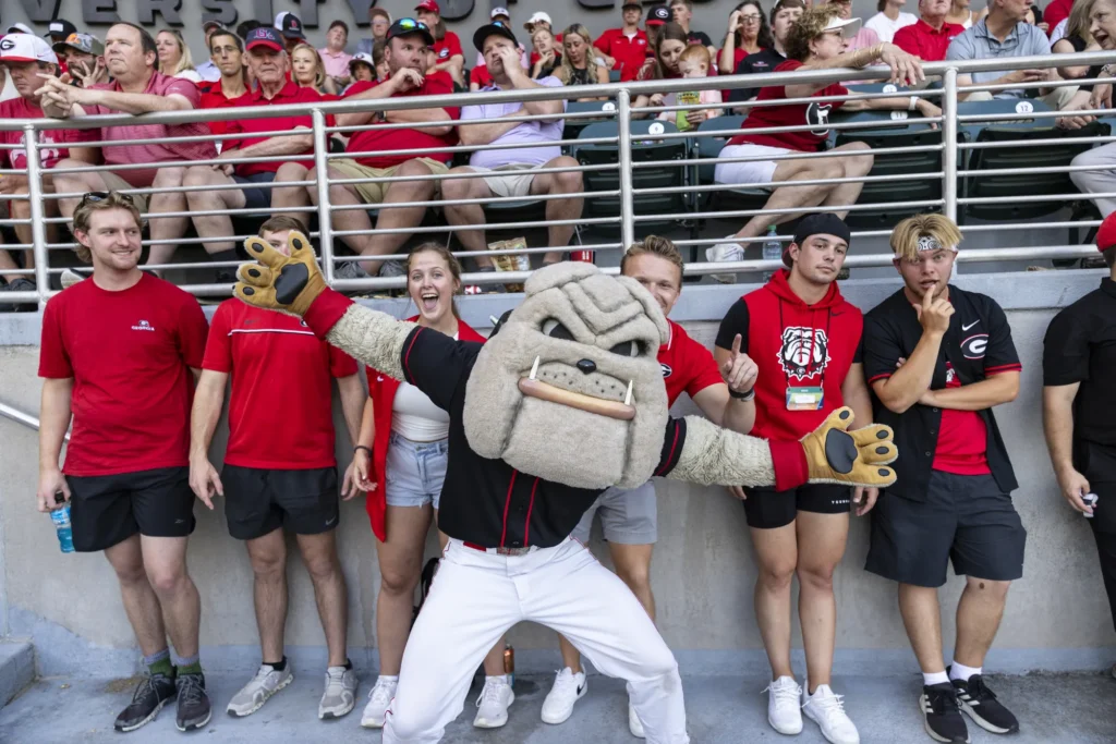 Hairy Dawg poses on the sidelines in a black shirt and white pants while UGA fans pose behind him.