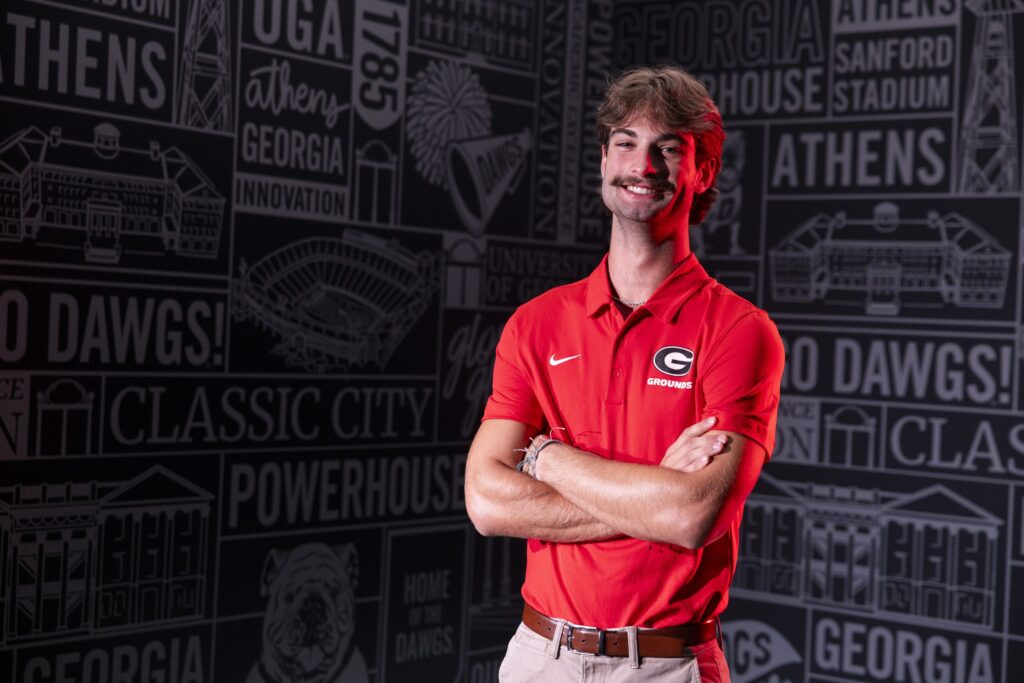 Tristan Lowery wears a red shirt with the UGA logo. He is standing in front of a black and white wall with UGA related text and images.