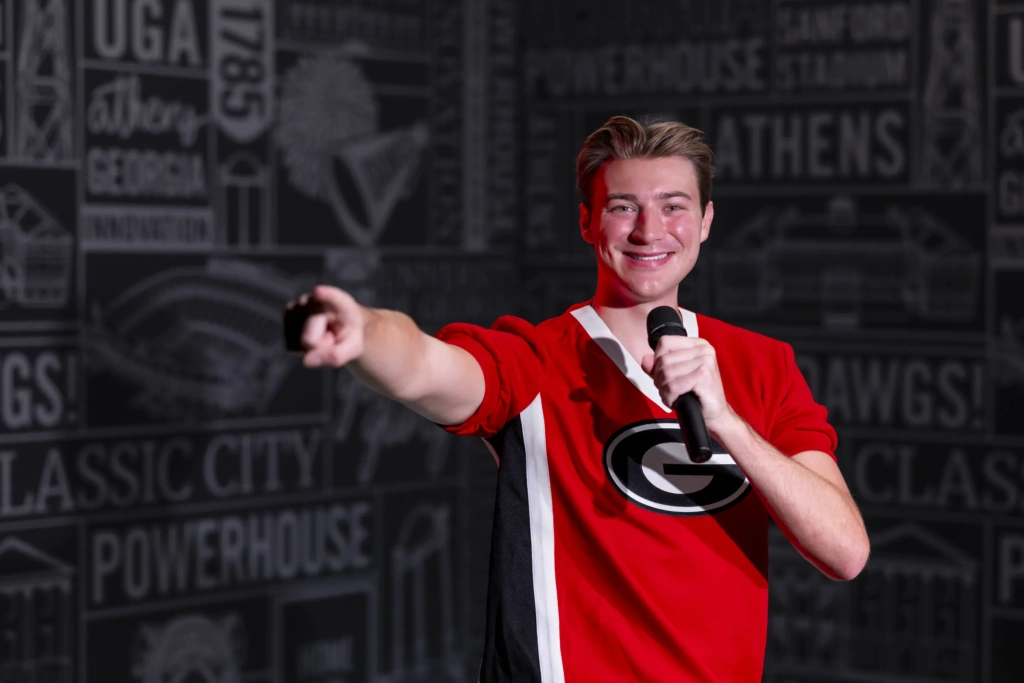 A young man wearing a red Georgia Bulldogs shirt holds a microphone toward his mouth while his other hand points toward the viewer.