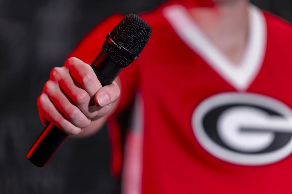 A close-up detail shot of a hand holding a microphone in the foreground. The person holding that microphone wears a red Georgia Bulldogs shirt in the background.