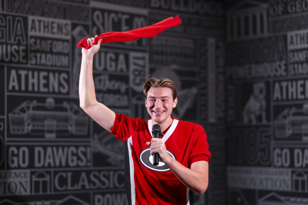 A young man wearing a red Georgia Bulldogs shirt swings a red towel over his head and holds a microphone to his mouth.