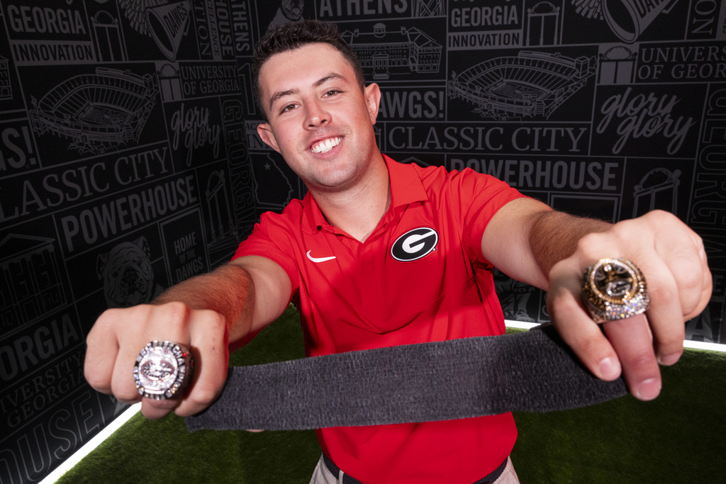 A young man in a red polo is wearing a championship ring on one finger of each hand. He holds a strip of black medical tape.