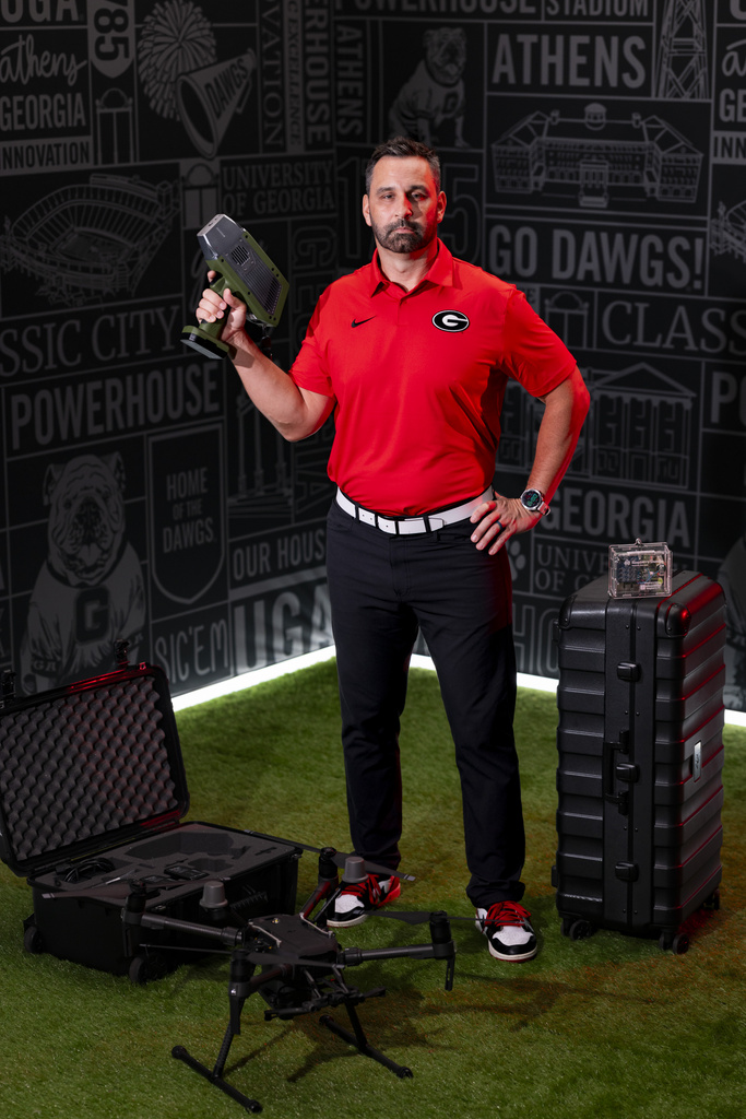 A man wearing a red polo and black shirt stands next to a tall, black tool case while holding a green and black power tool.