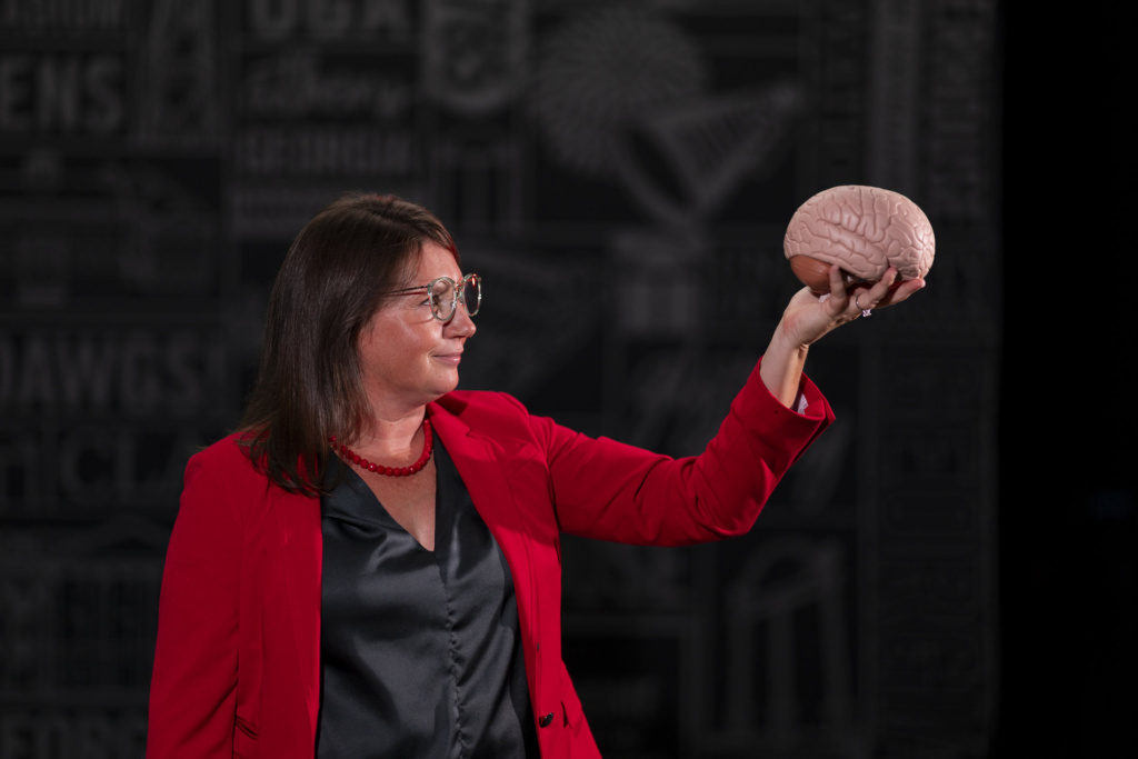 A woman wearing glasses and a red blazer holds up a life-sized plastic brain model in her raised hand against a dark backdrop.