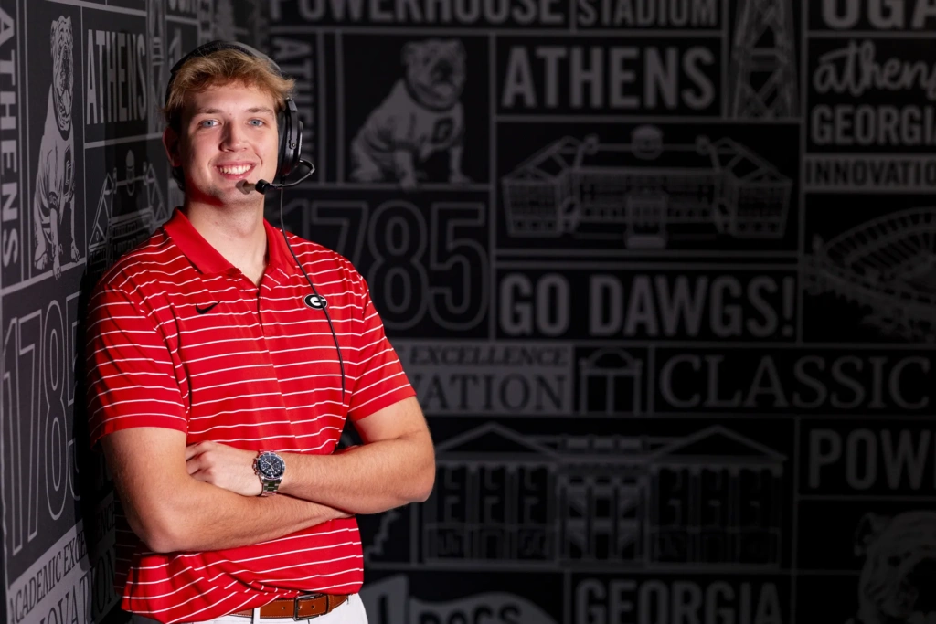 A young man wearing a red and white striped polo and a black headset stands against a dark wall with his arms crossed.