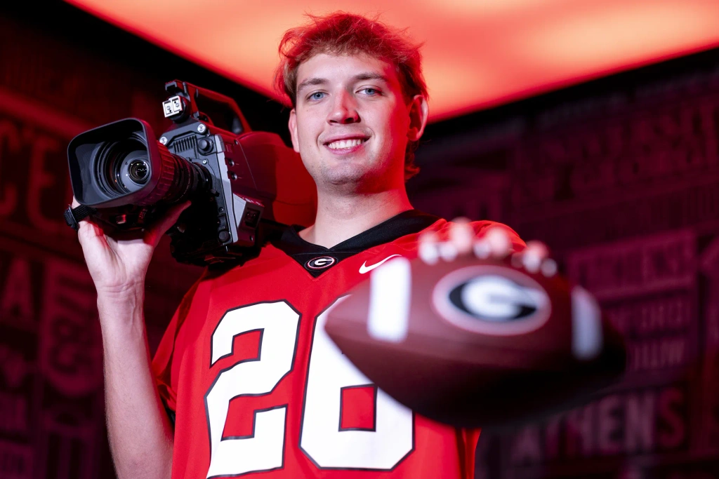 A closeup of a young man wearing a Georgia Bulldogs jersey and holding a blurred football in one hand and a large camera on the opposite shoulder.