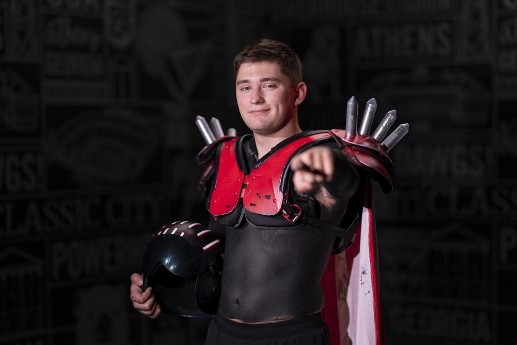 A young man smiles and points at the camera. He wears red, spiked shoulder pads and a long cape. His torso is painted black and he is holding a pilot's helmet under one arm.