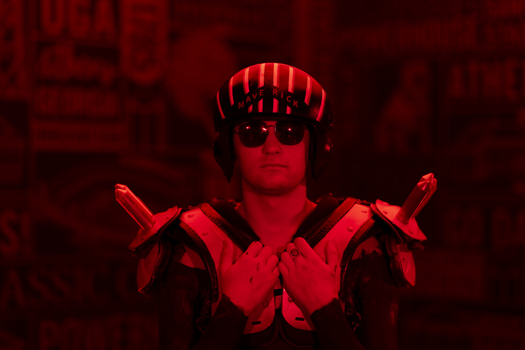 A close up of a young man wearing a pilot's hemet, sunglasses, and spiked shoulder pads. The room is dimly lit with a red light.
