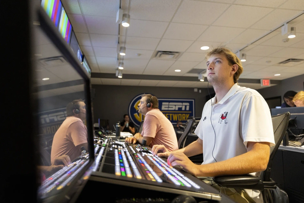 A young man wearing a white polo shirt operates a brightly lit sound board.