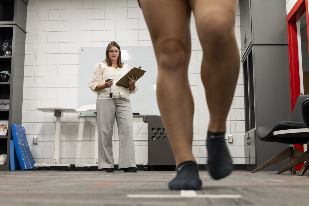 A closeup of a pair of legs walking on a gray floor as a woman observes in the background.