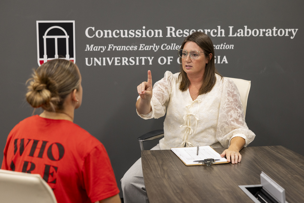 A woman wearing a white blouse holds up an index finger in front of another woman whose back is to the camera. They are in UGA's Concussion Research Laboratory.